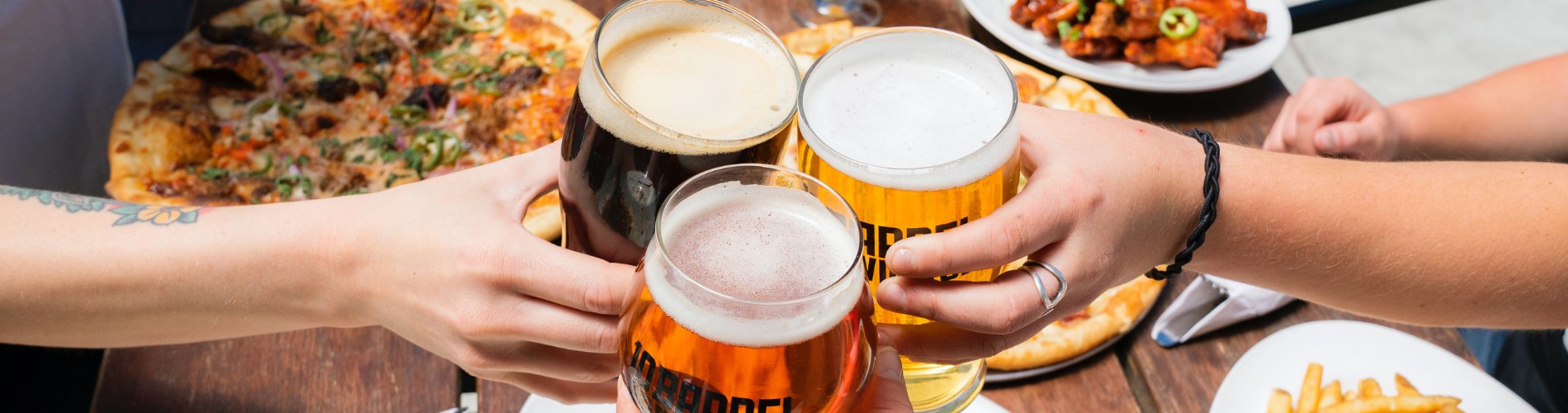 Three people holding pints of beer over a table of pizzas