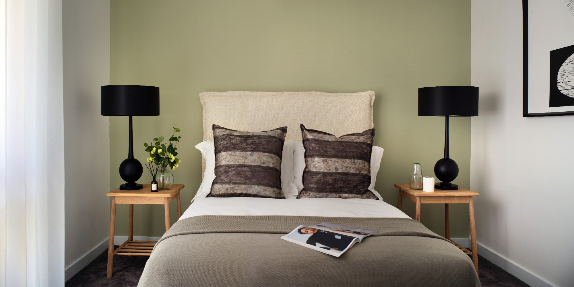 A double bed against a green wall with neutral toned cushions and duvet, with an open magazine on the bed